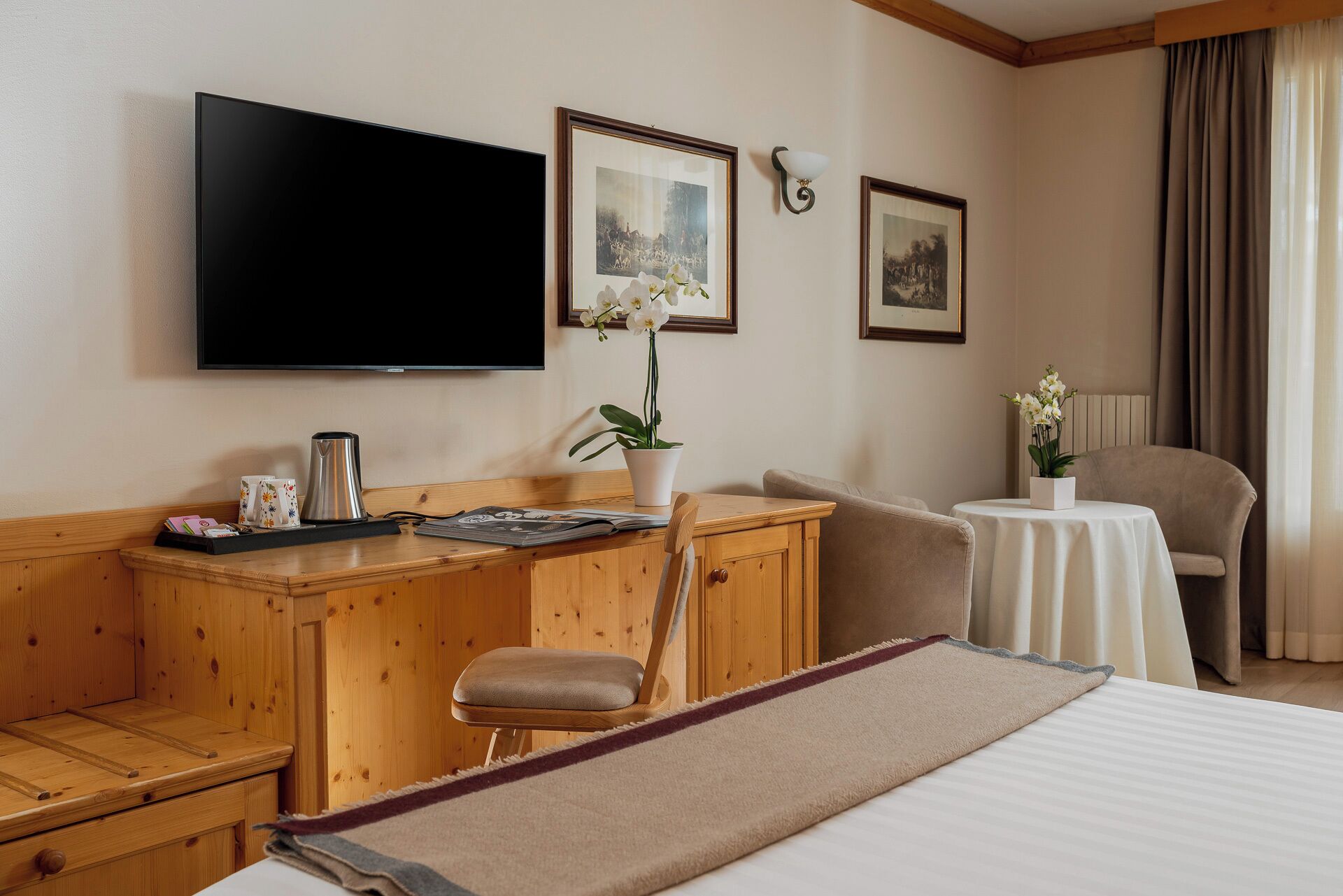 A hotel room with a wooden desk, a television, a bed, a chair, and a small table with a vase of flowers.