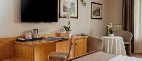 A hotel room with a wooden desk, a television, a bed, a chair, and a small table with a vase of flowers.