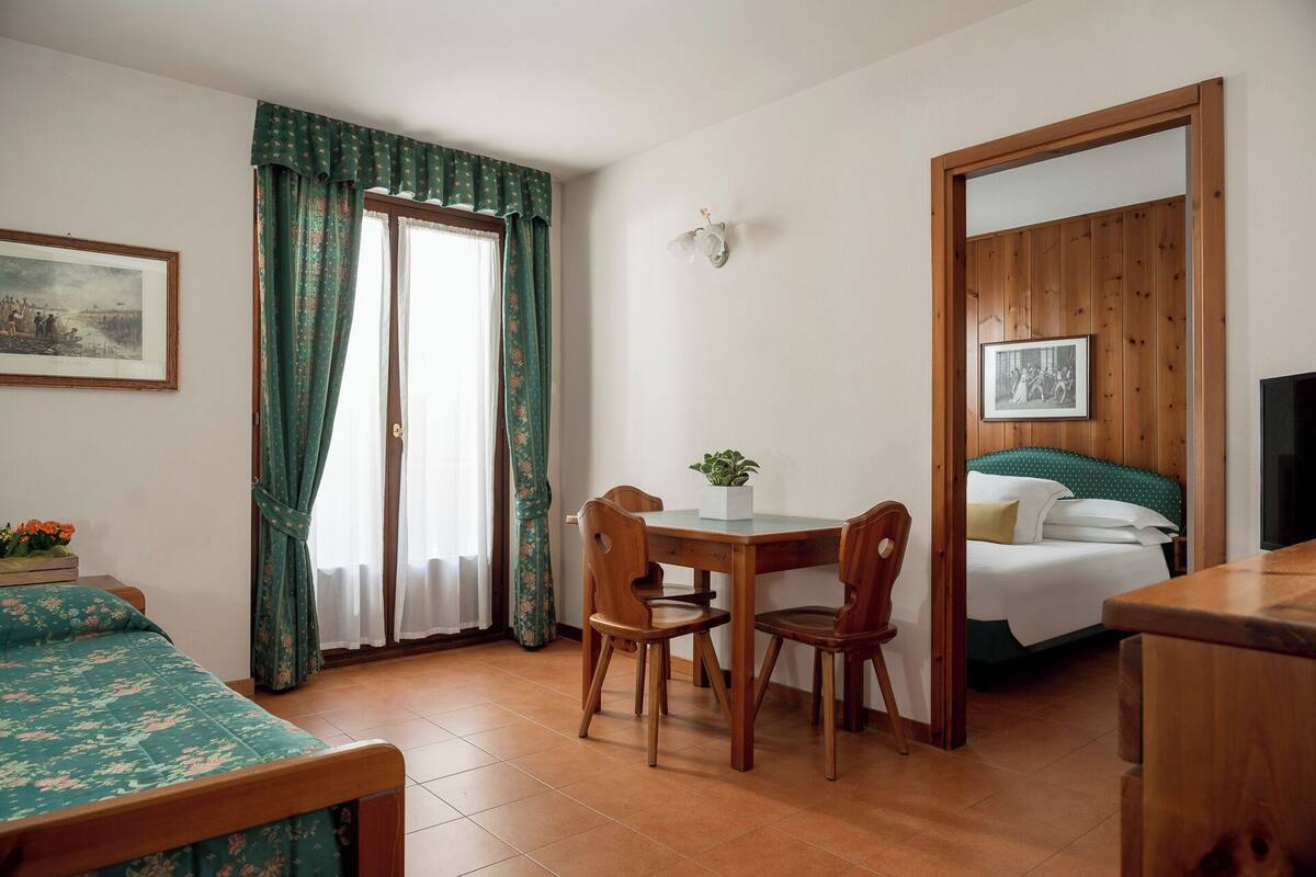 A room with a bed, a desk, two chairs, a small table with a plant, and a framed picture on the wall.