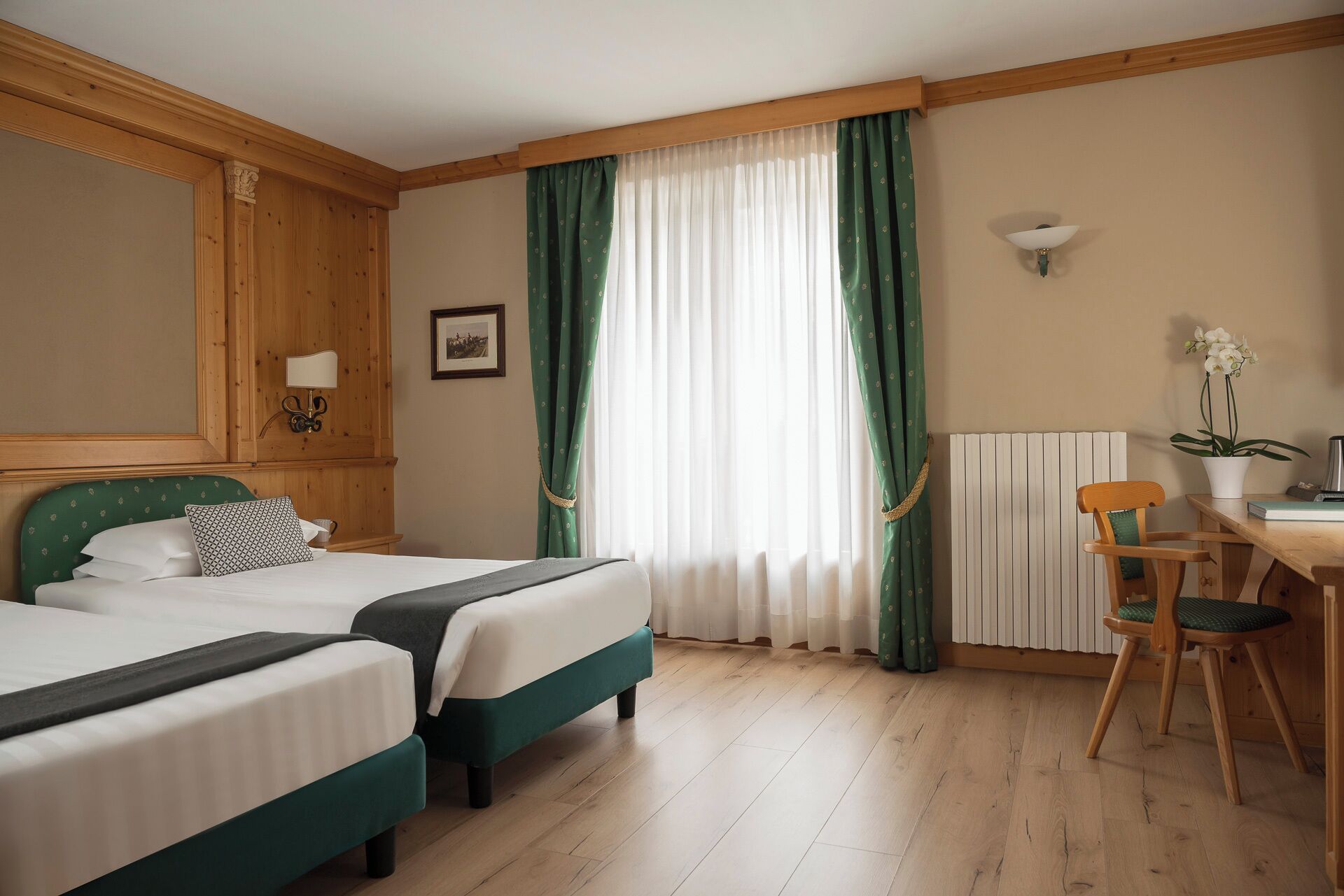 A hotel room with two beds, a desk with a chair, a radiator, and a picture on the wall.