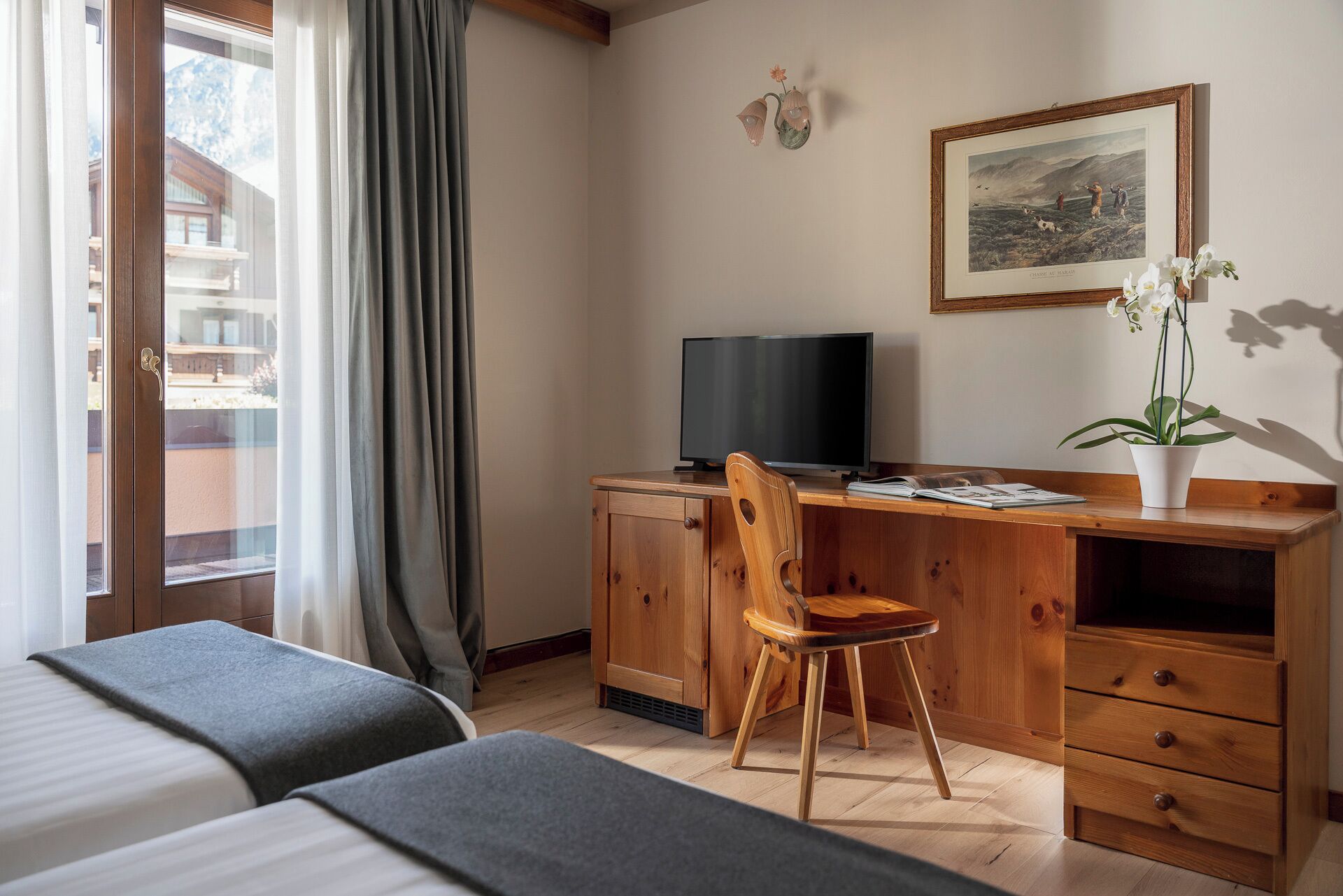 A hotel room with a wooden desk, a television, a chair, a bed, and a window with curtains.
