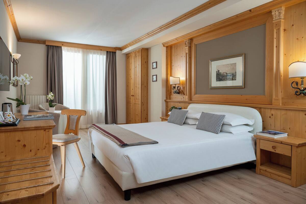 A hotel room with a large bed, wooden headboard, a desk with a chair, a bedside table with a lamp, and a framed picture on the wall.