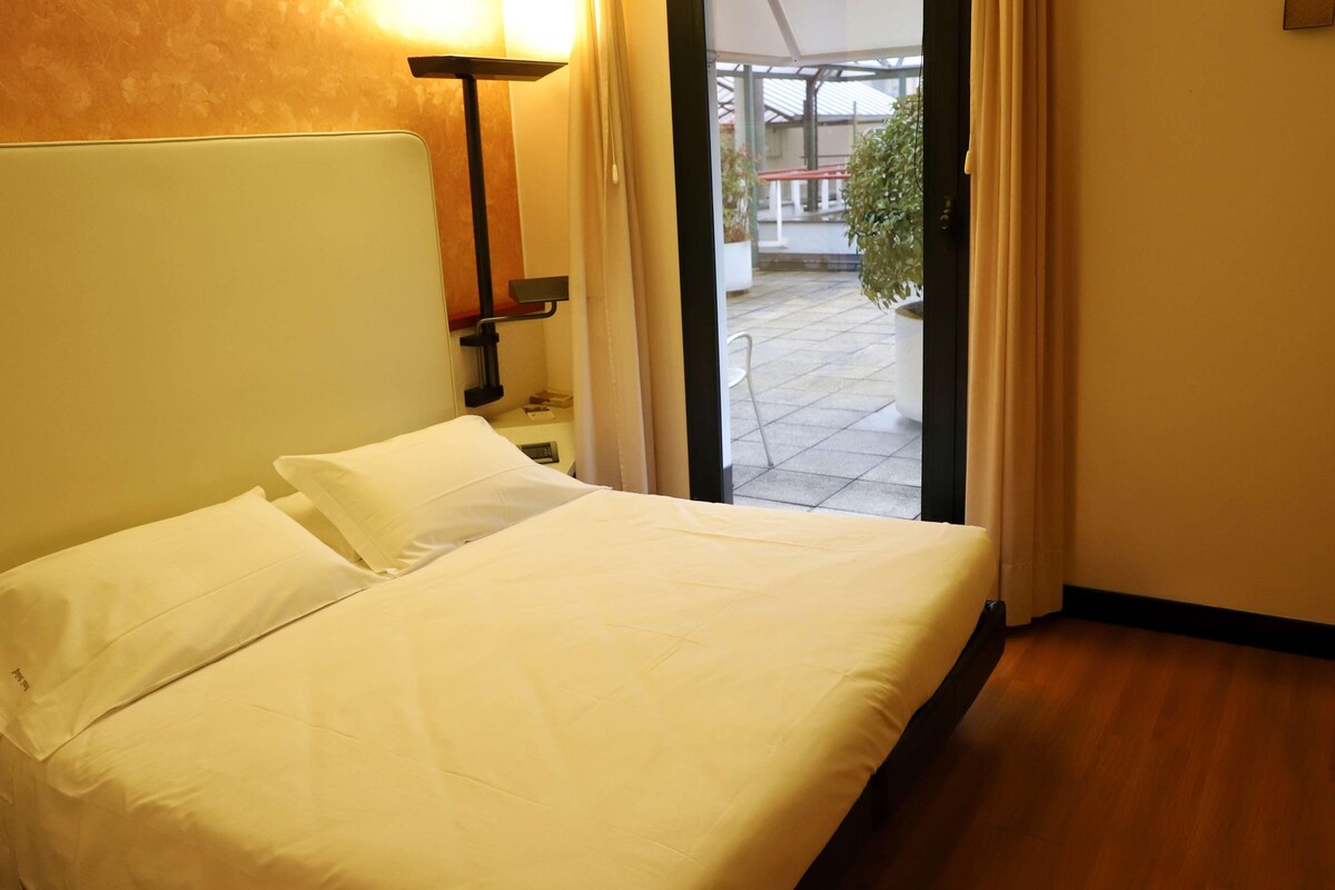 A hotel room with a bed, a nightstand, a lamp, and a view of the outdoor area.