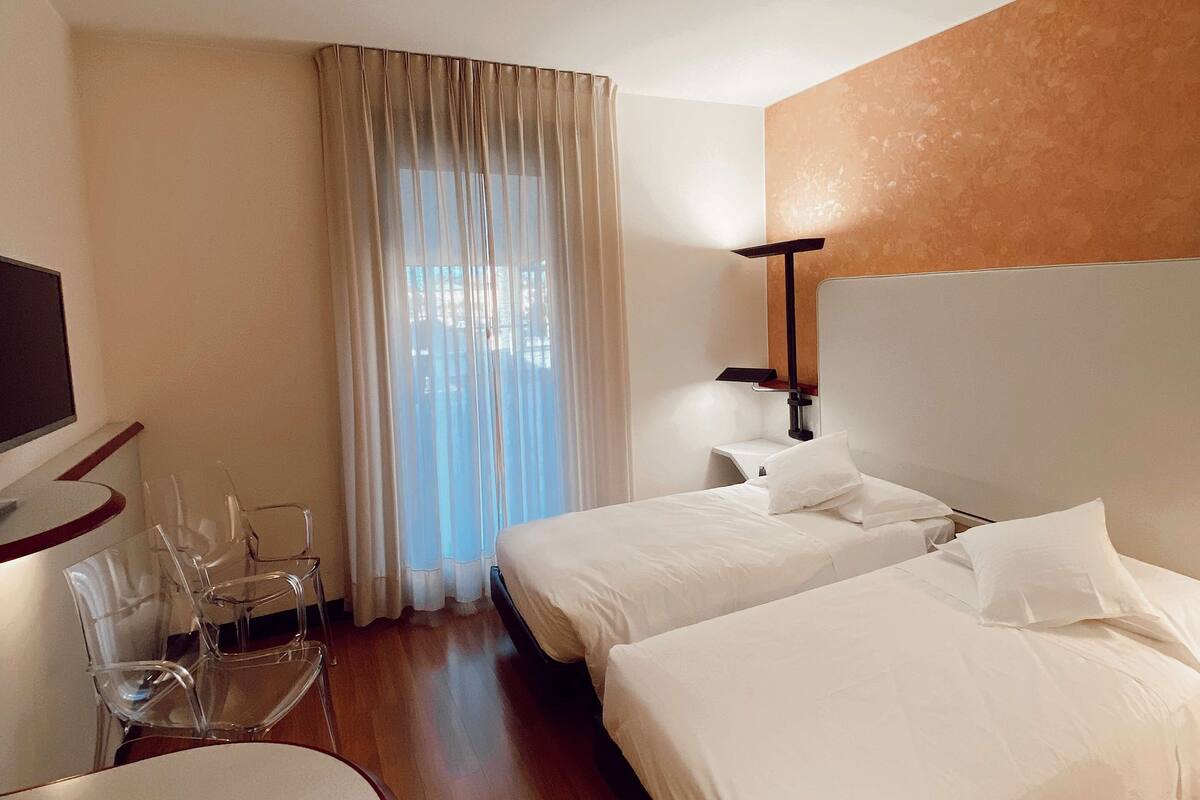 A hotel room with two beds, a TV, a desk with chairs, and a window with curtains.