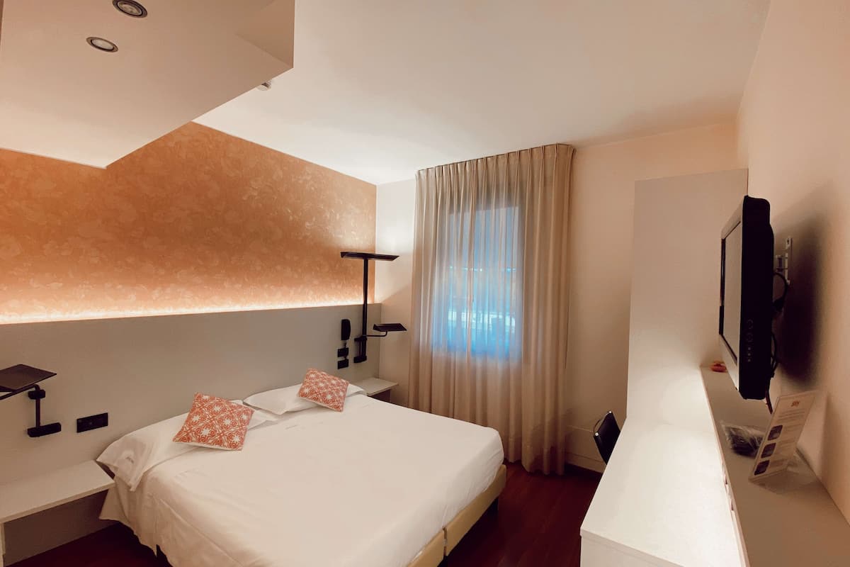 A modern hotel room with a large bed, a desk, a TV, and a window with curtains.