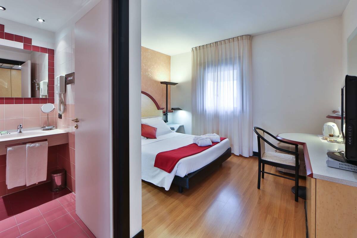 A hotel room with a bed, bedside table, lamp, TV, and a small desk with a chair.