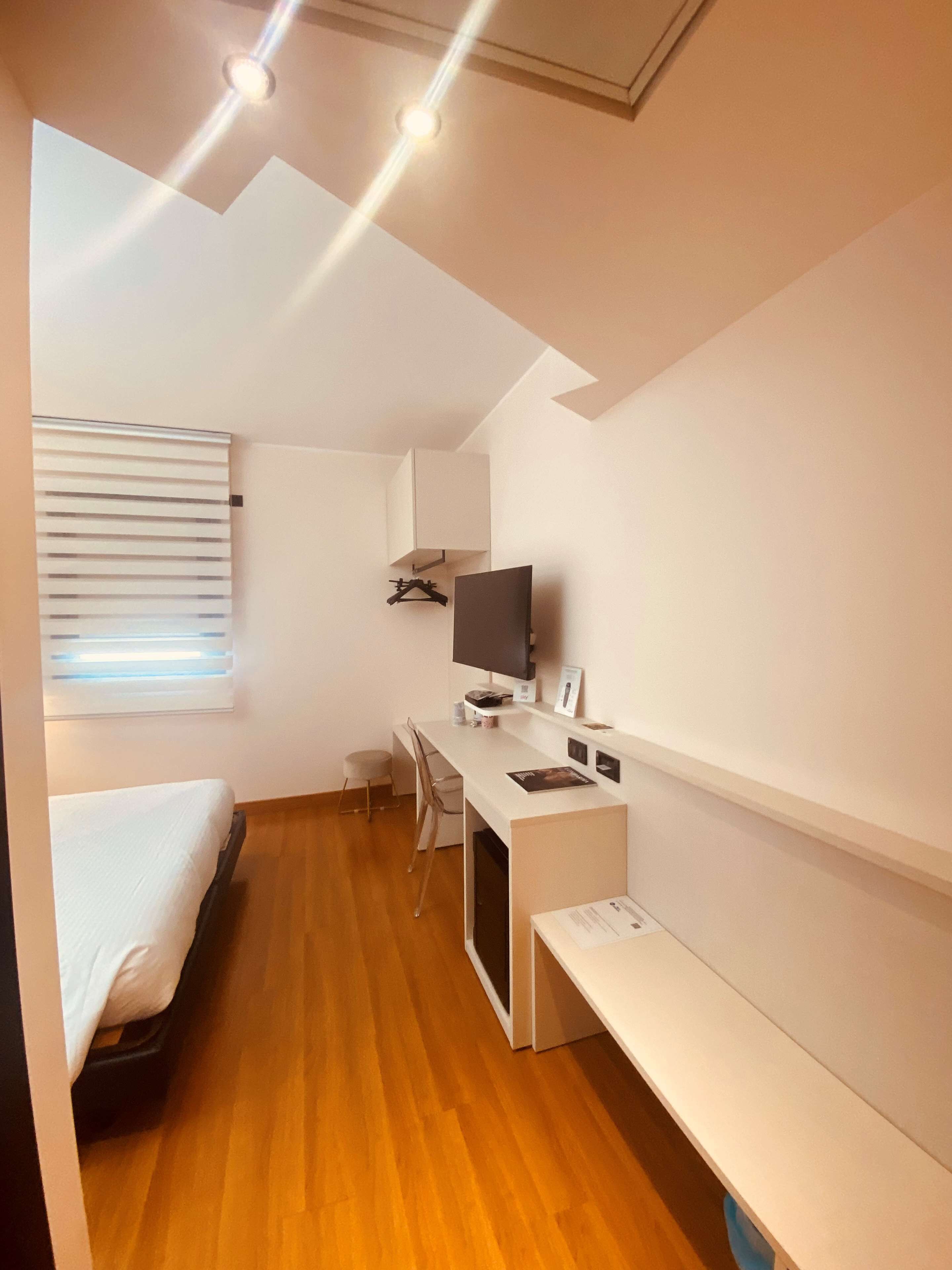 A compact hotel room with a bed, a desk, a chair, a TV, and a small window with blinds.