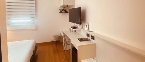 A compact hotel room with a bed, a desk, a chair, a TV, and a small window with blinds.