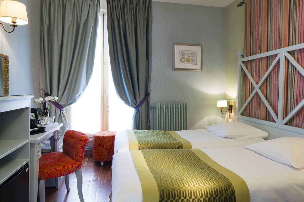Una camera d'albergo con due letti, una scrivania con una sedia, un termosifone e una finestra con tende.