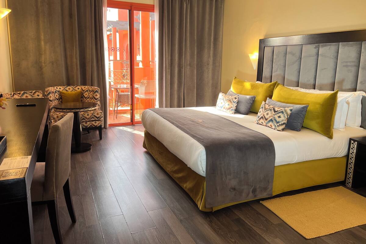 A hotel room with a bed, a desk, and a chair. There is a sliding glass door leading to an outdoor area with orange pillars.