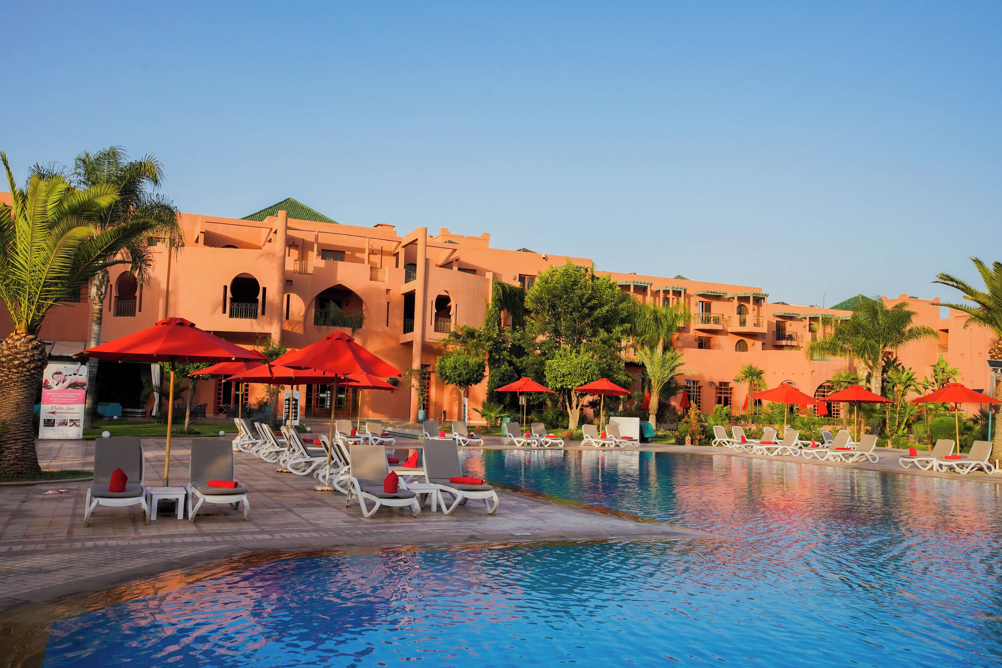 A pool area with lounge chairs and red umbrellas, surrounded by a building with multiple balconies and arches.