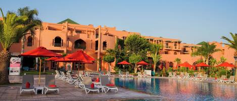 A pool area with lounge chairs and red umbrellas, surrounded by a building with multiple balconies and arches.