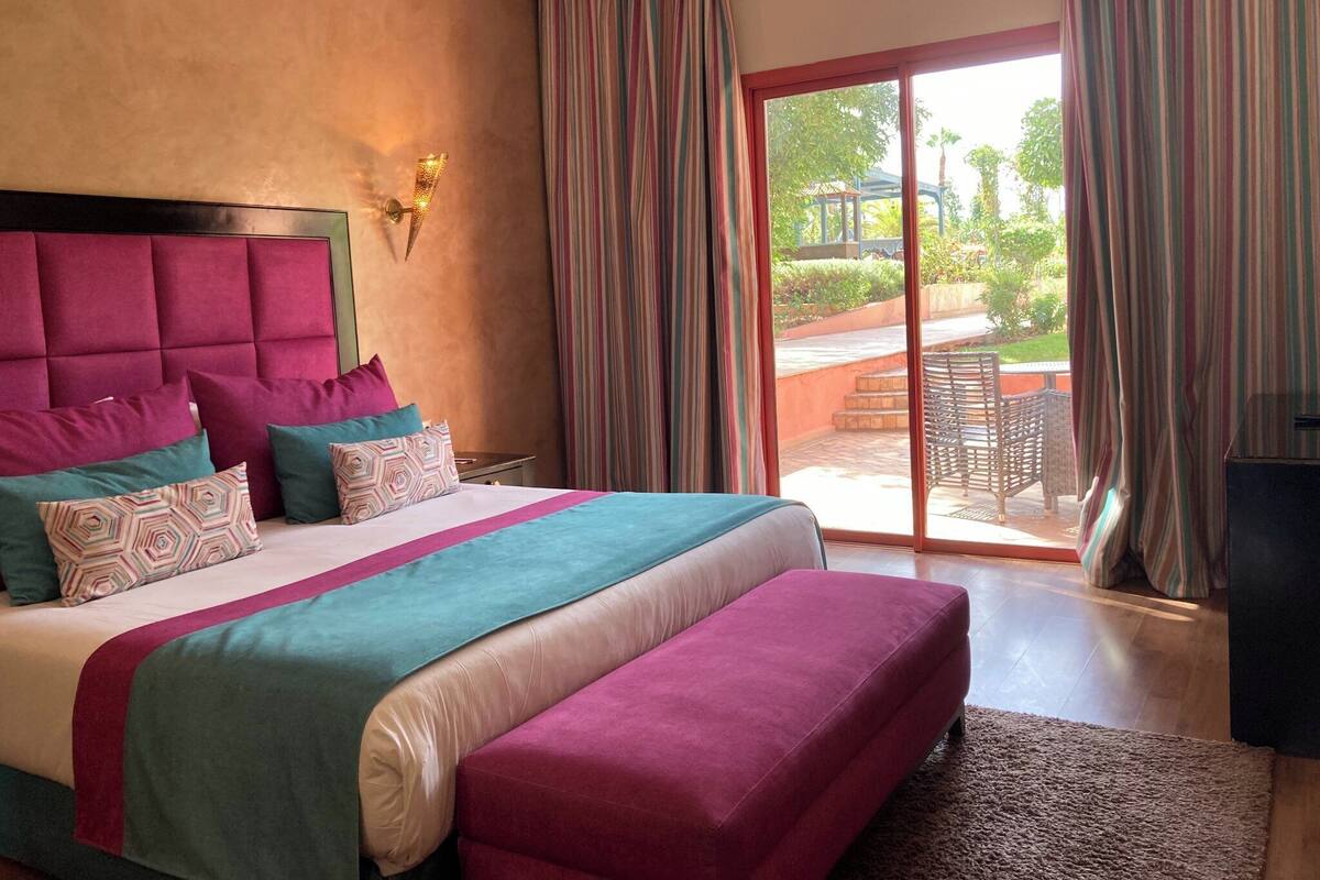 A hotel room with a large bed, a headboard, a bedside table, a bench, and a view of an outdoor area with a patio and chairs.