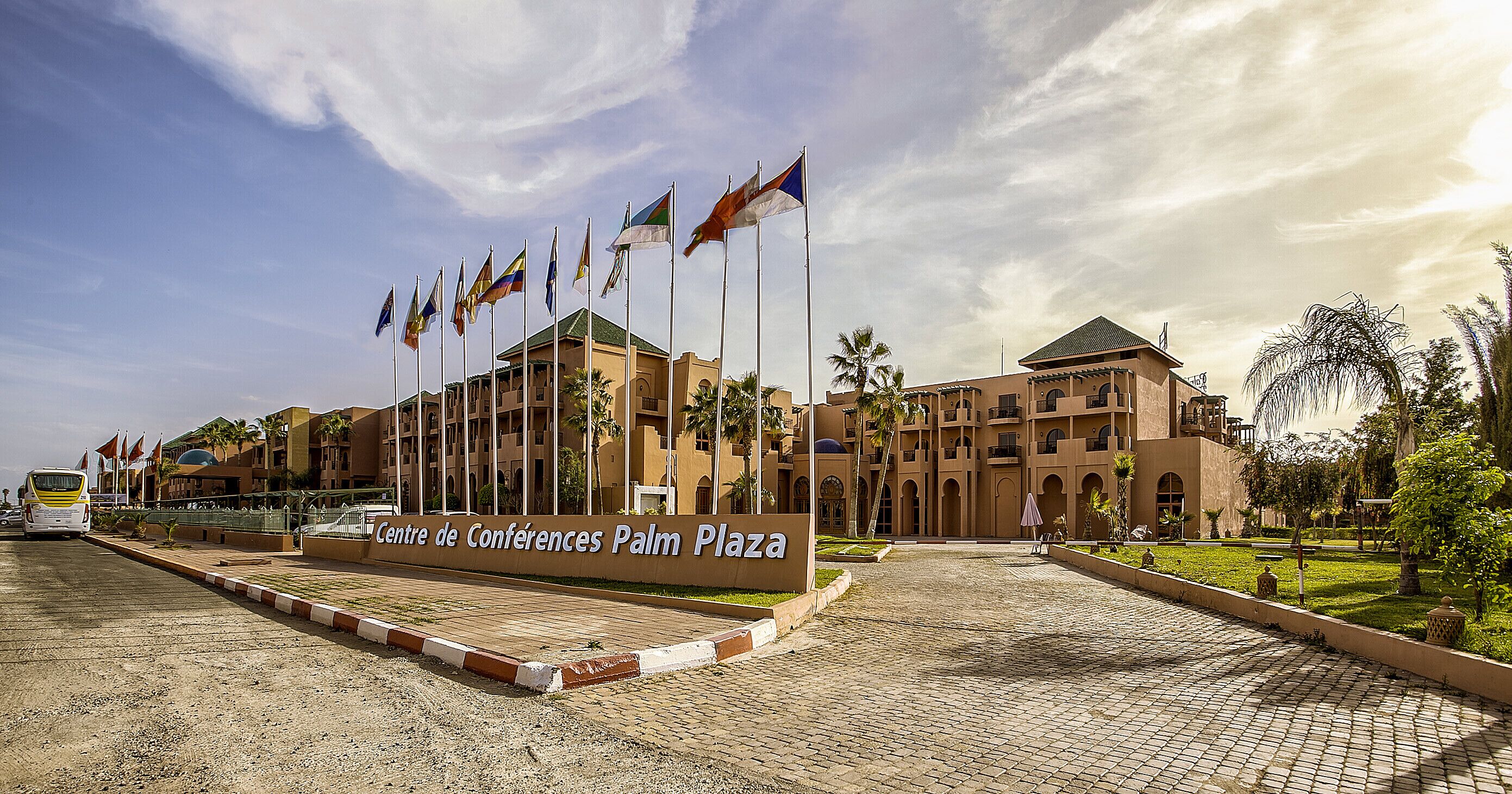 A building with multiple flags, a sign reading "Centre de Conferences Palm Plaza", and a paved entrance area with palm trees.