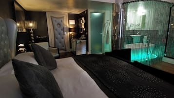 A modern hotel room with a large bed, a sitting area, a small kitchenette, and a bathroom with a glass shower.