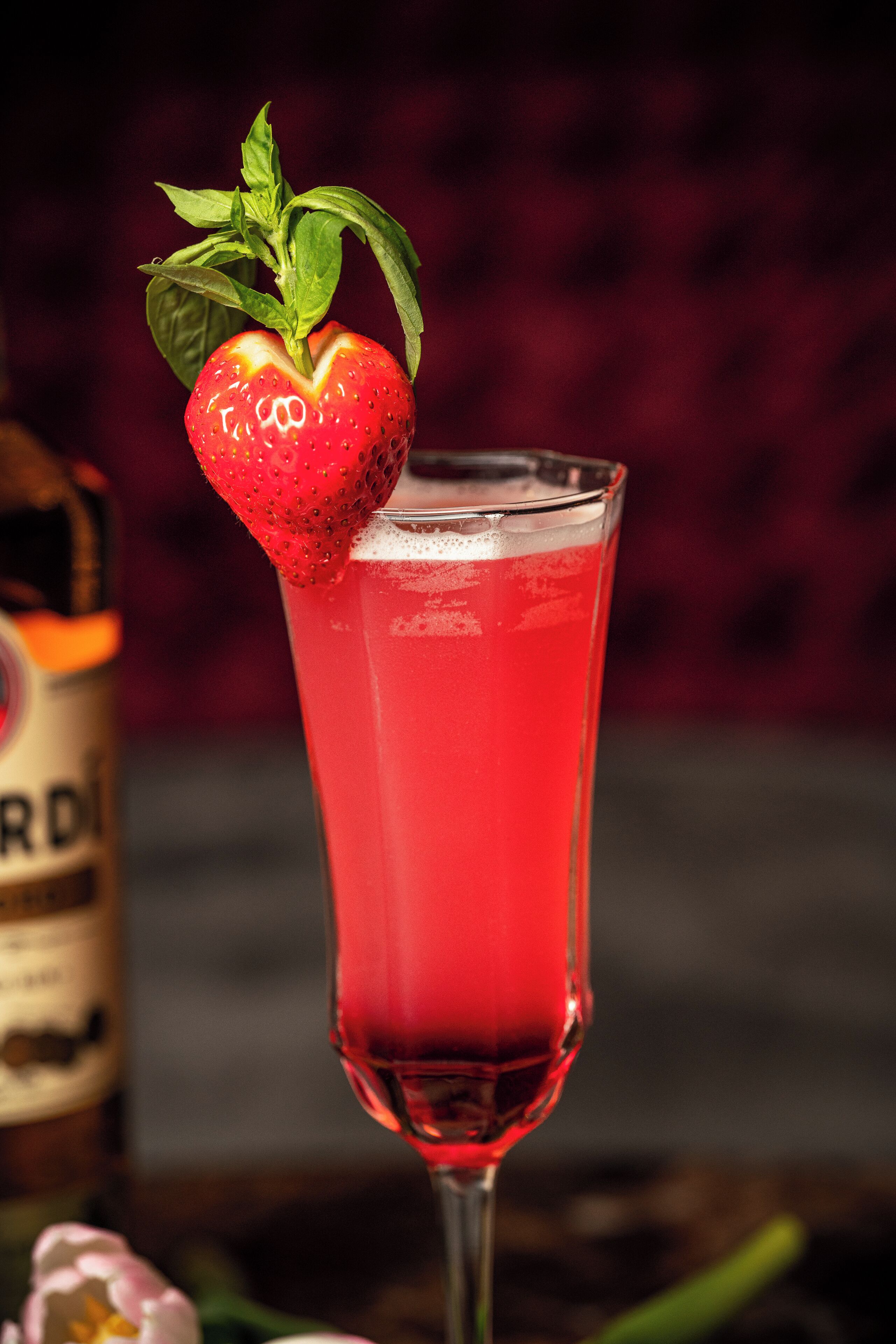 Un grand verre contenant une boisson rouge, décoré d’une fraise, et une bouteille en arrière-plan.