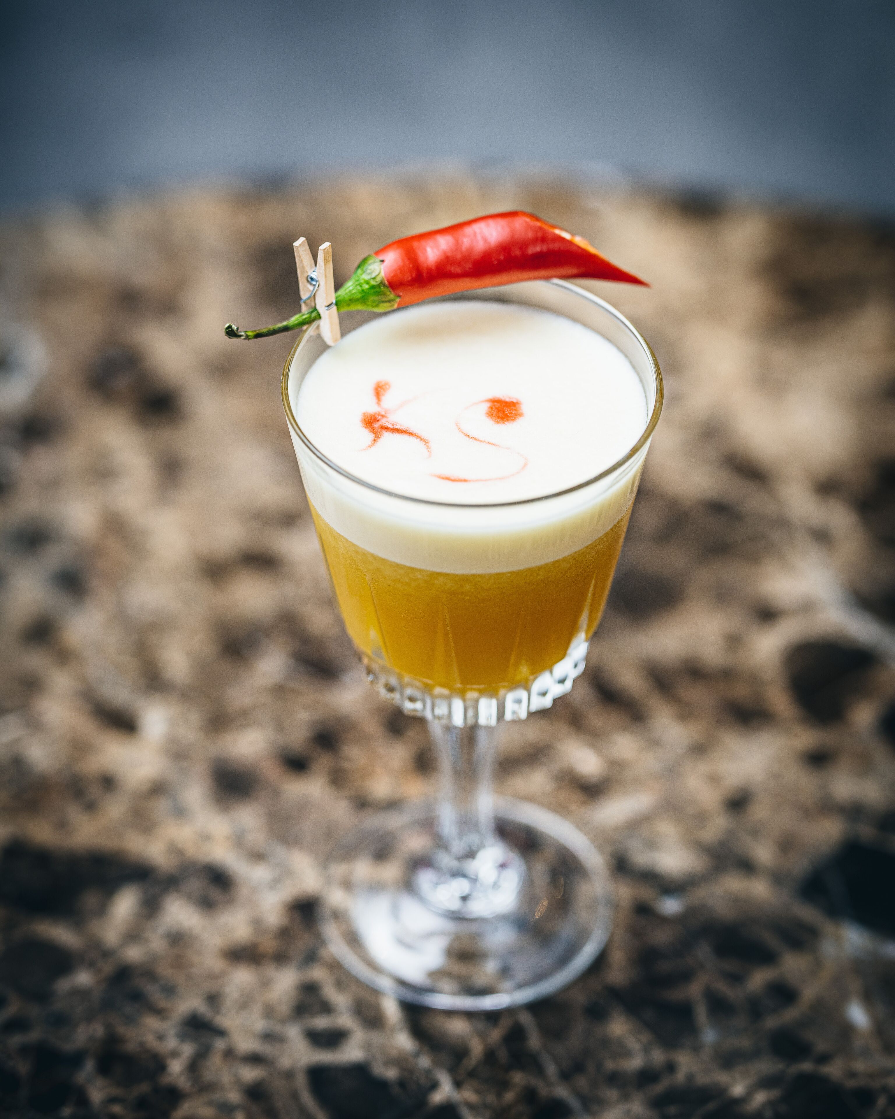 Un cocktail servi dans un verre à martini, garni d’un piment rouge, le tout posé sur une surface texturée.