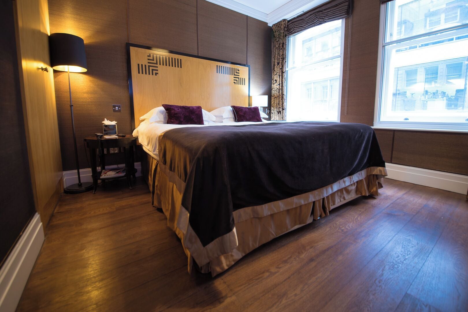 Ein ordentlich eingerichtetes Hotelzimmer mit einem großen Bett, einem Nachttisch, einer Lampe und Blick auf die Stadt durch ein Fenster.