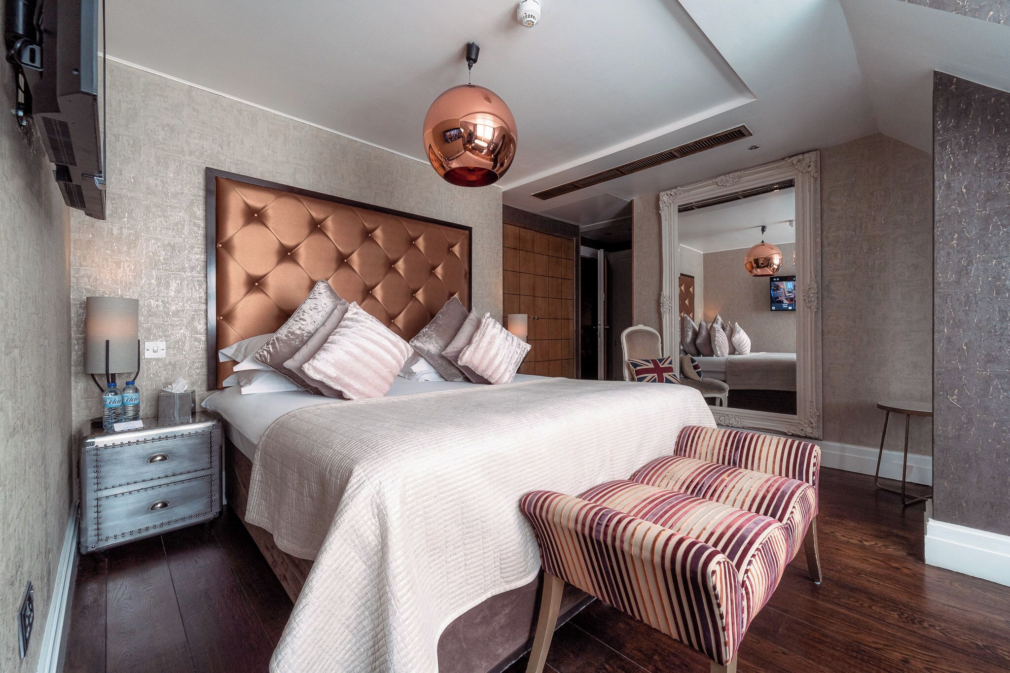 A modern bedroom with a tufted headboard, a large bed, a nightstand with drawers, a striped bench, and a mirror.