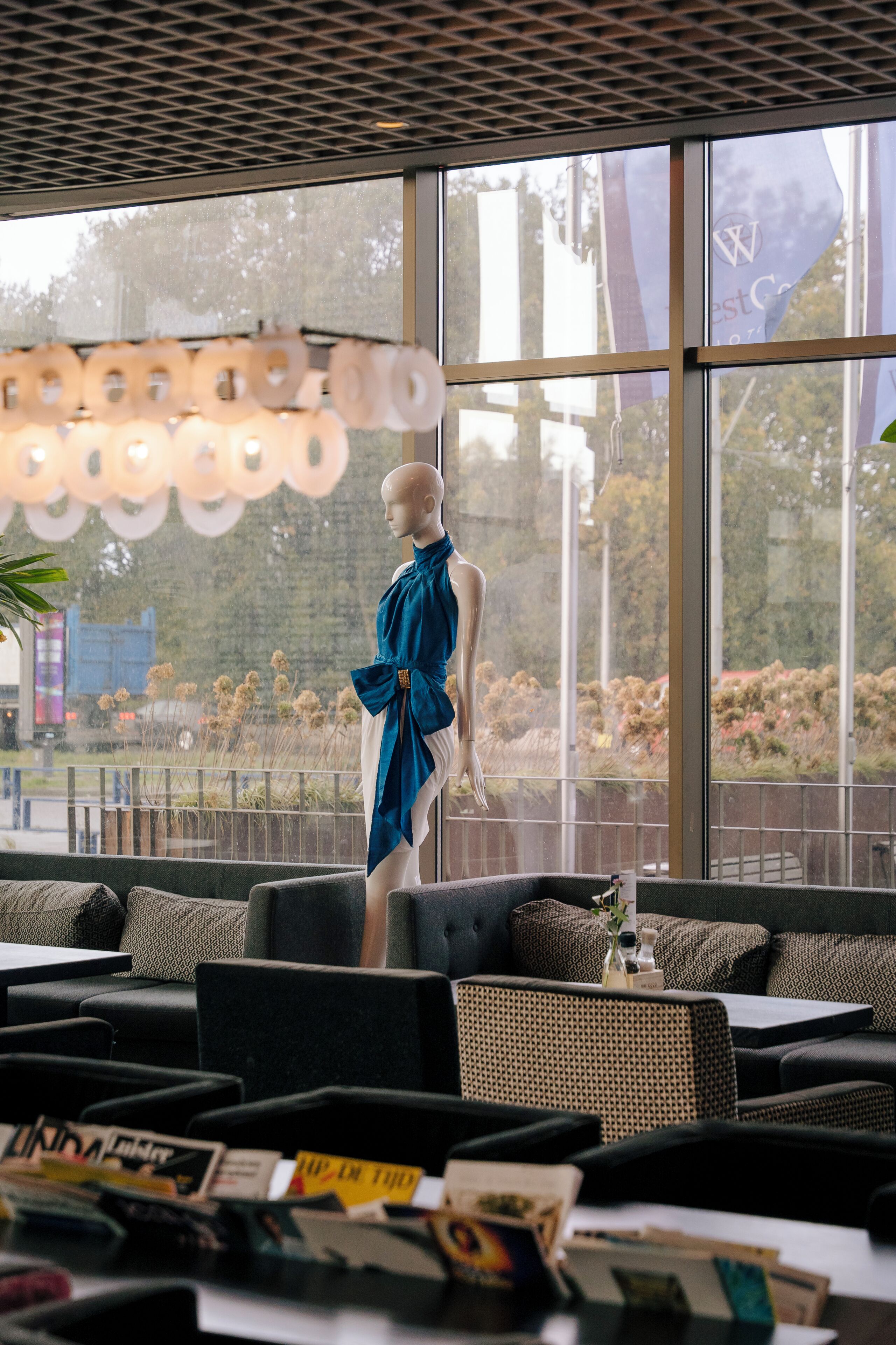 A mannequin in a blue dress standing by a large window with a view of trees and a fence.
