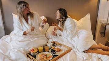 Dos personas en una habitación de hotel, sentadas en la cama con una bandeja de alimentos para el desayuno, incluyendo huevos, panqueques y pasteles.