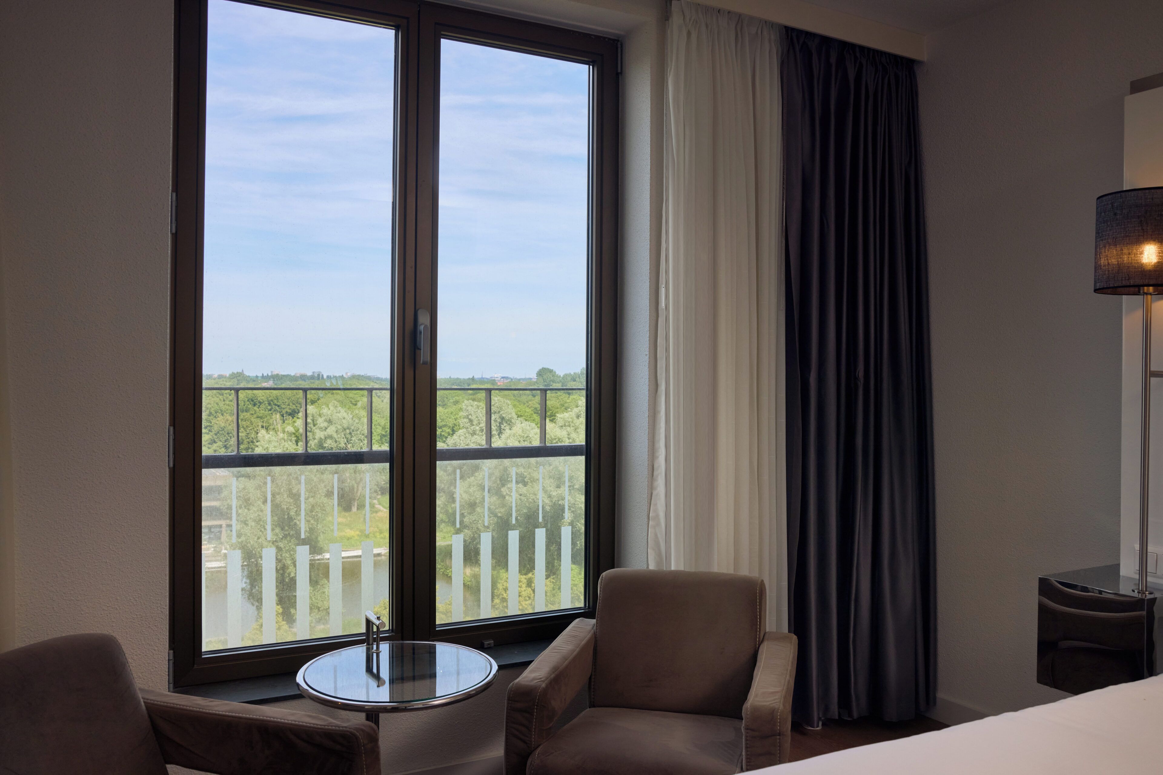 A hotel room with a large window, a round table, two chairs, a bed, and a lamp.
