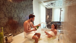 Un hombre y una mujer en una bañera con champán, en un baño moderno con papel tapiz estampado y cabina de ducha de vidrio.