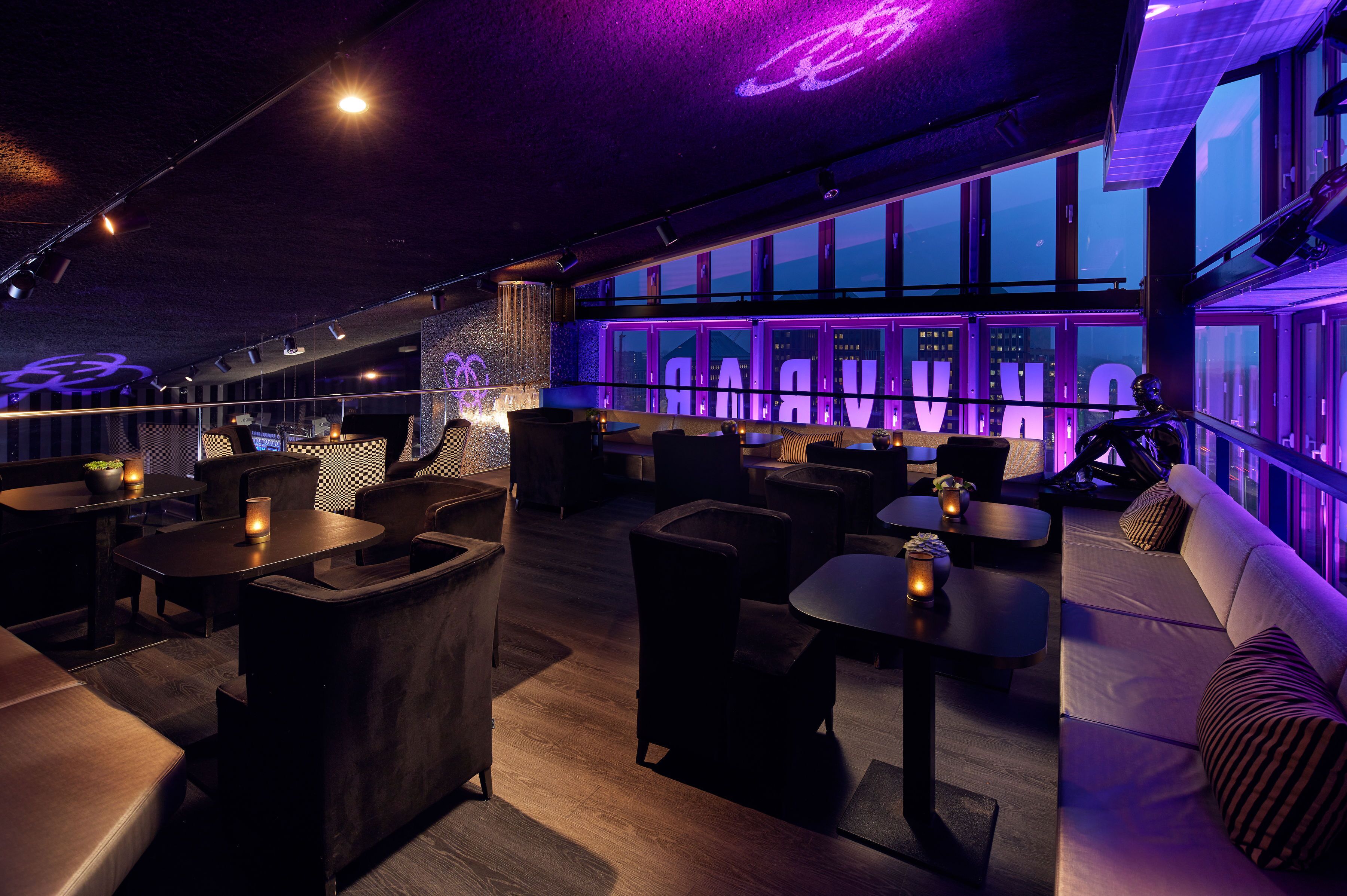 A modern lounge area with seating arrangements, a bar, and large windows offering a city view.