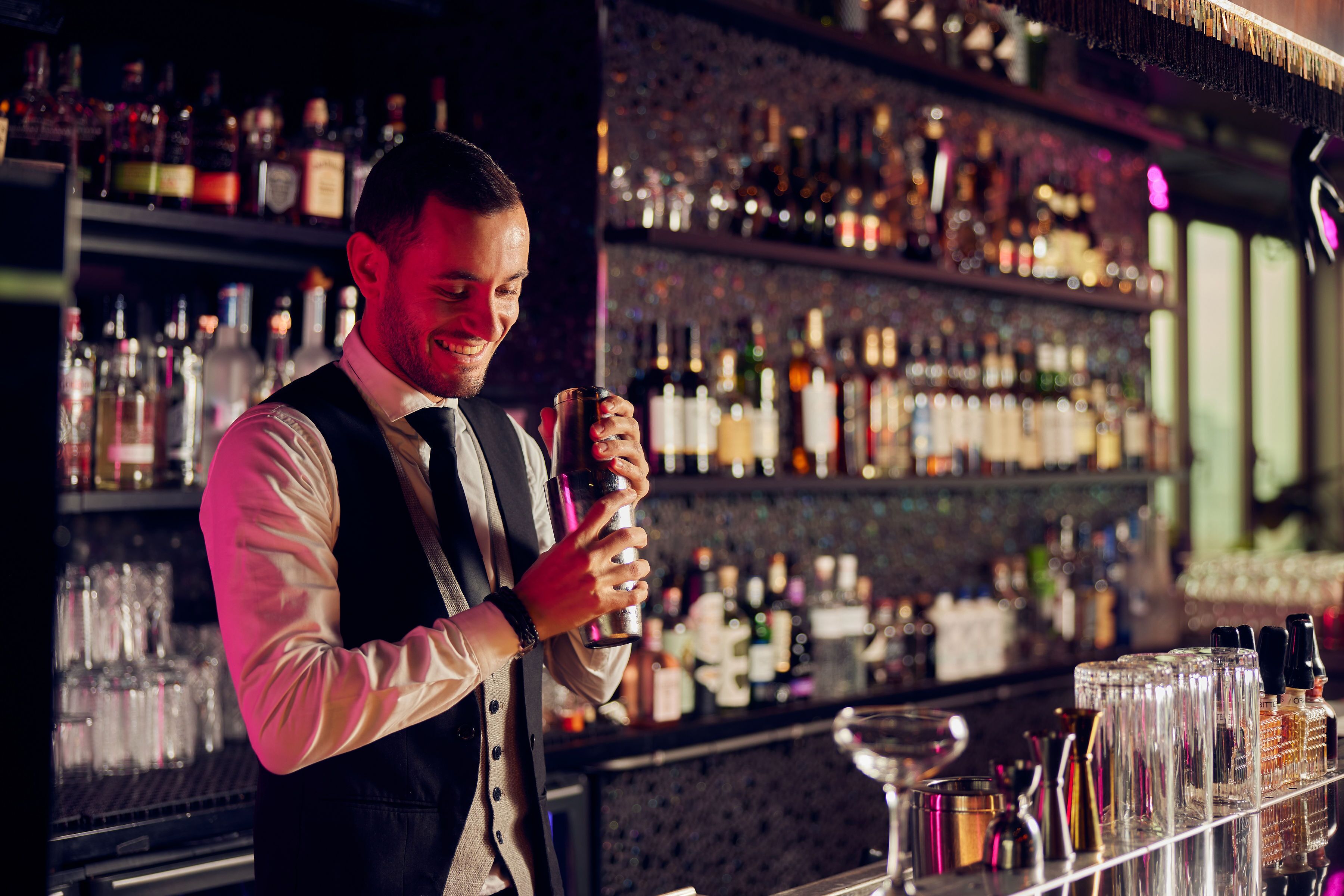 Un barman derri ère un comptoir où l’on peut voir des étagères remplies de bouteilles et de verres de toutes sortes.