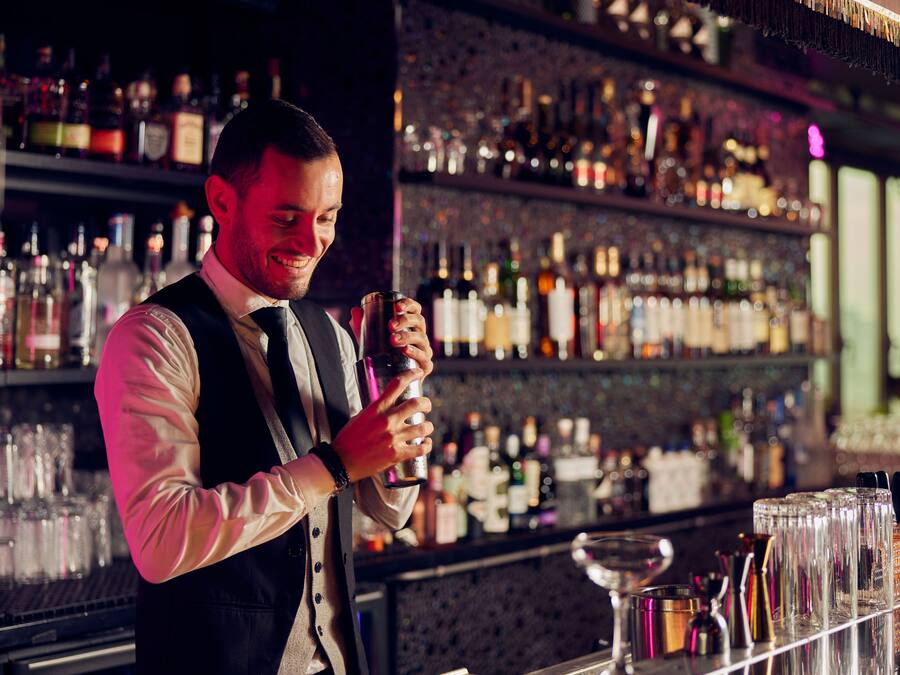 Un cantinero detrás de una barra con estantes llenos de botellas y vasos.