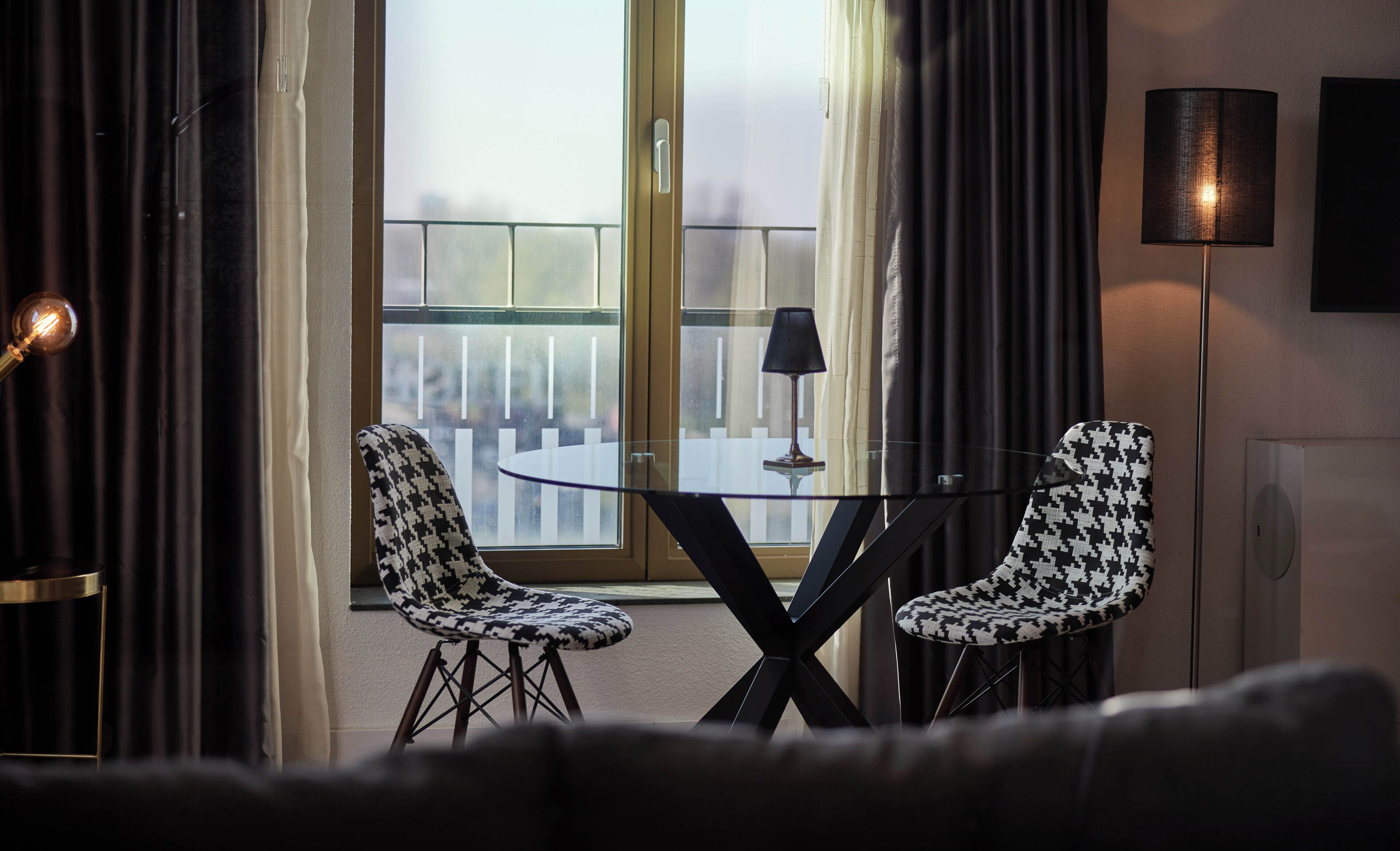 Une pièce moderne avec une table en verre et deux chaises à motifs, une lampe et une vue sur l’extérieur à travers une fenêtre avec des rideaux.