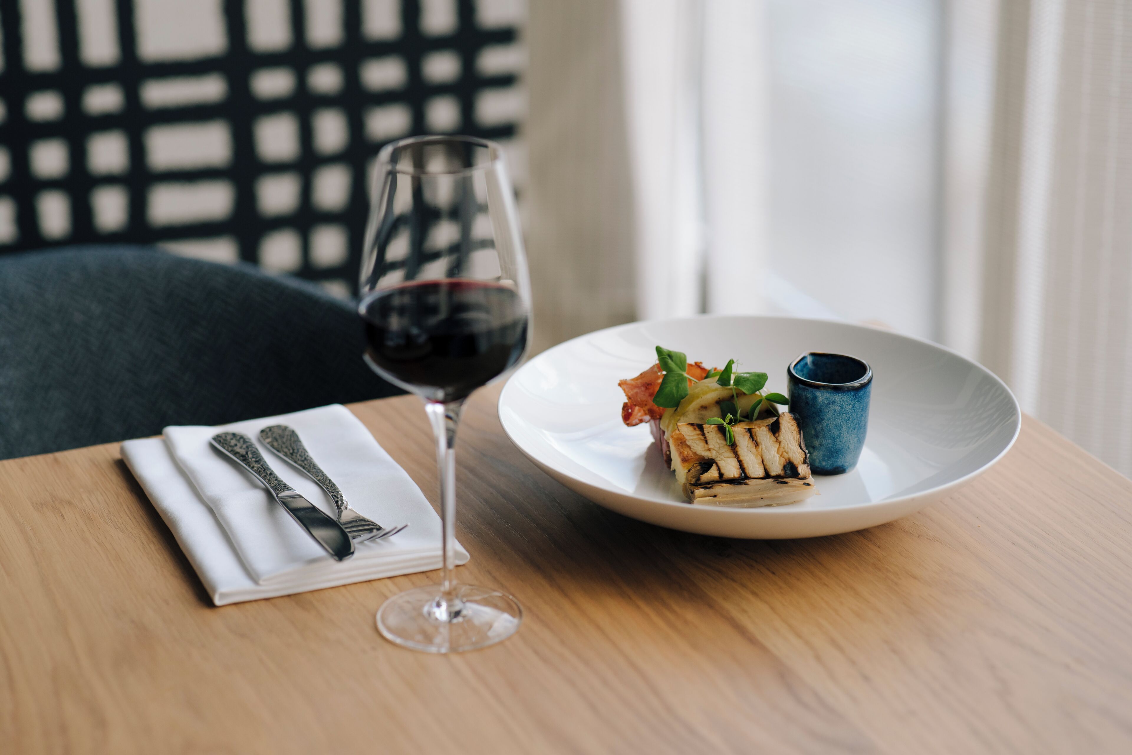 A dining table with a white plate containing a grilled dish, a small blue cup, and a glass of red wine.