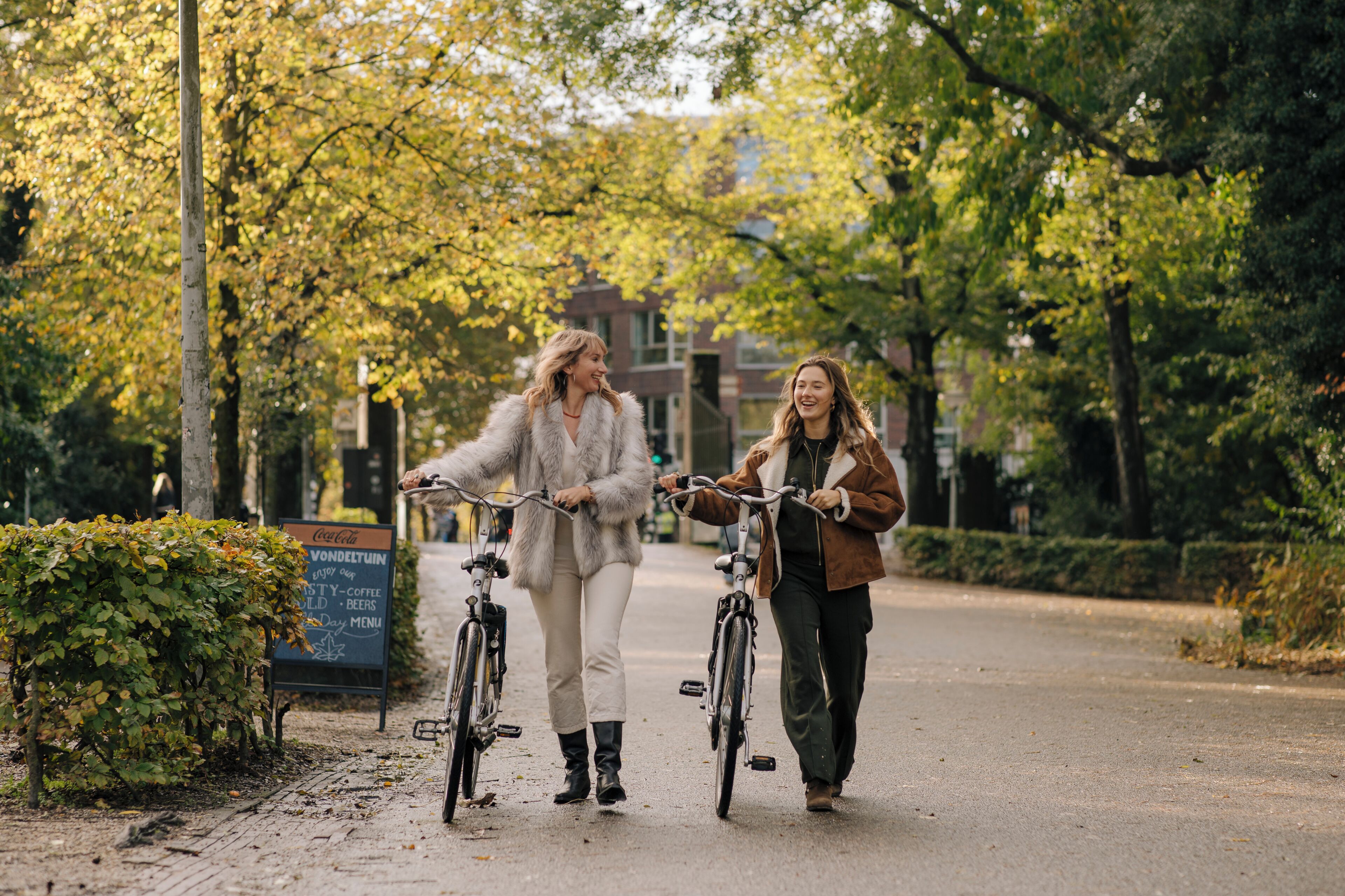 Two individuals walking with bicycles on a paved path, surrounded by trees and a signboard.