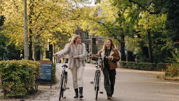 Two individuals walking with bicycles on a paved path, surrounded by trees and a signboard.