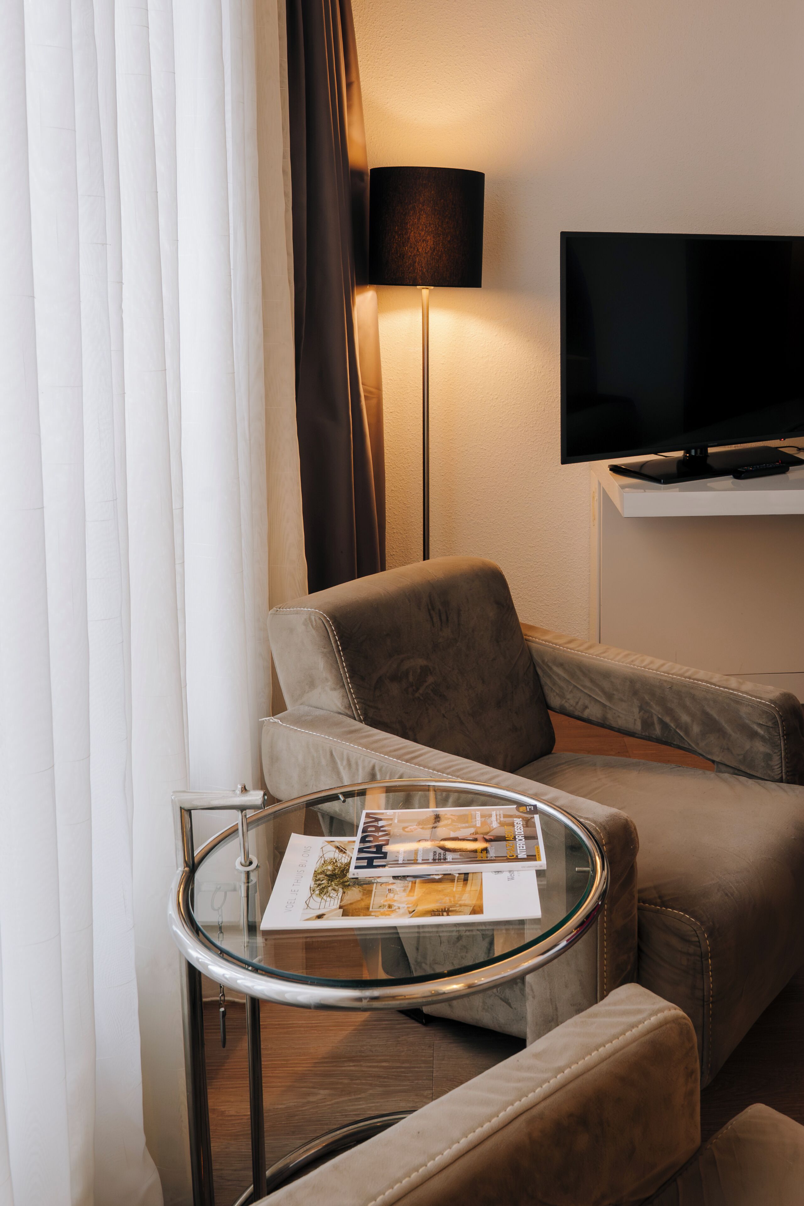 A modern hotel room with a glass coffee table, a sofa, a flat-screen TV mounted on the wall, and a lamp.