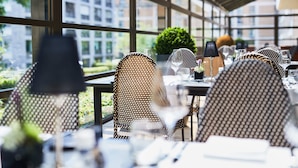 Un espacio exterior para comer con sillas y mesas de mimbre, ventanas amplias y vistas a edificios y zonas verdes.