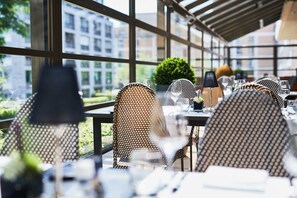 Een terras met rieten stoelen en tafels, grote ramen en uitzicht op gebouwen en groen.