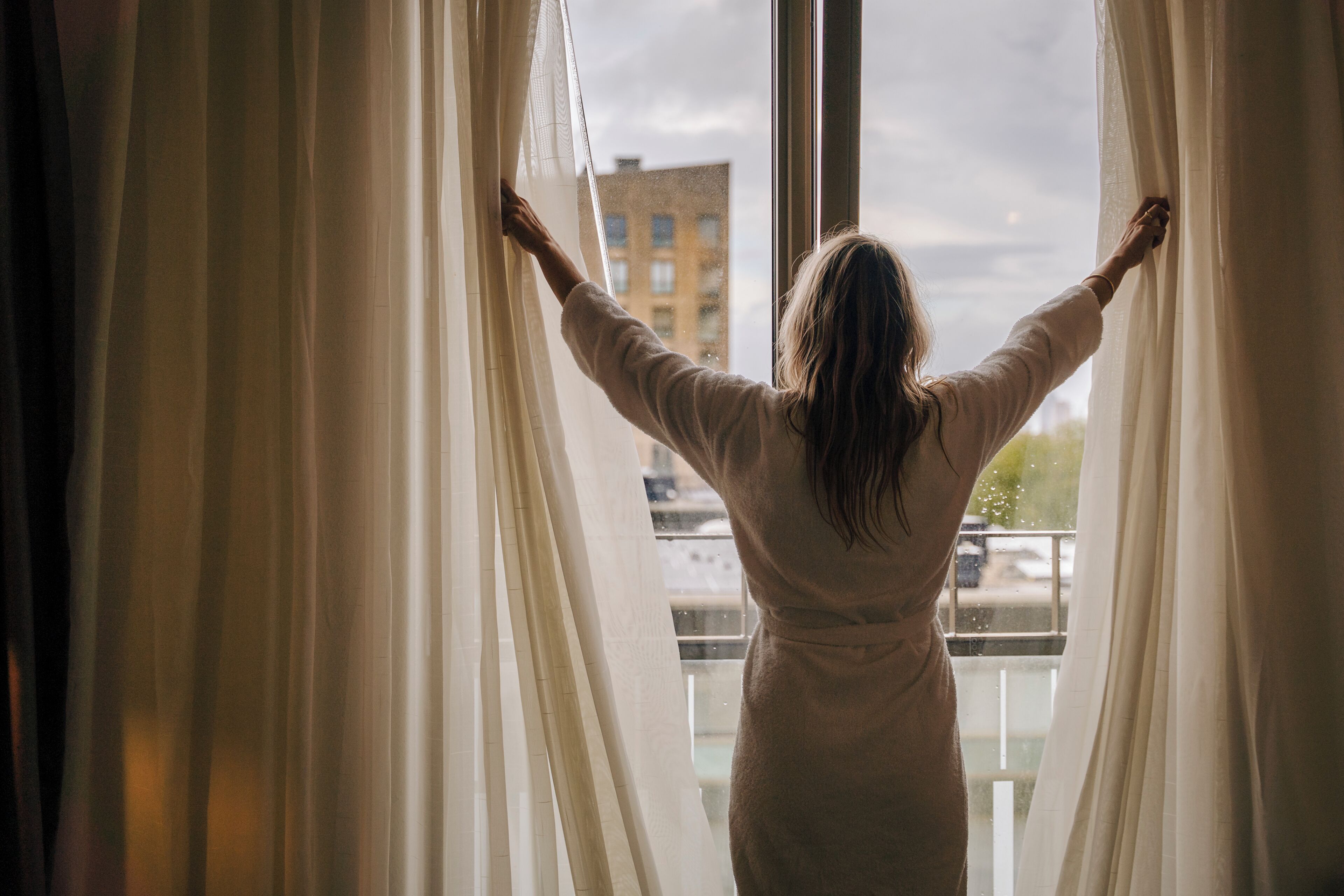 Una persona de pie junto a una ventana con cortinas translúcidas, con los brazos extendidos, mirando una vista urbana.