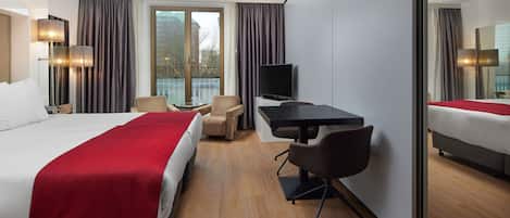 A modern hotel room with a large bed, a desk, a chair, a TV, and a balcony view.
