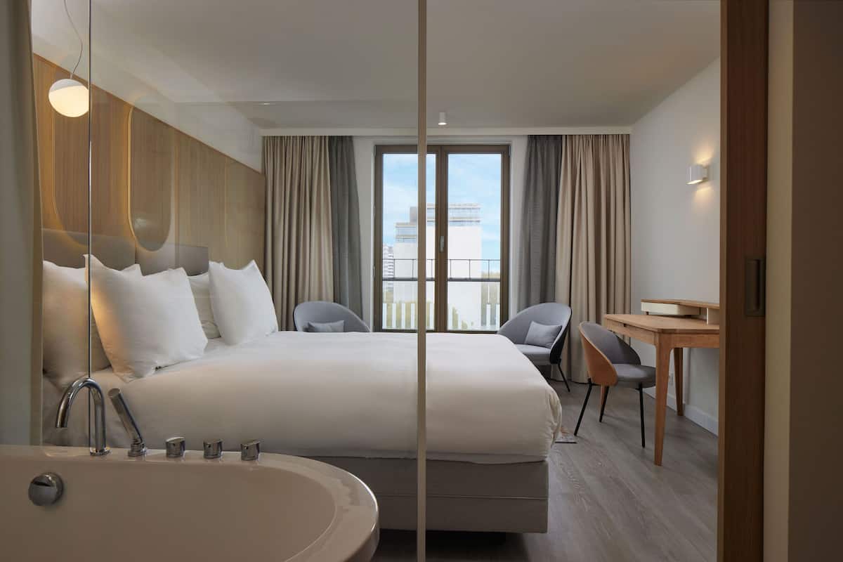 Een moderne hotelkamer met een groot bed, een bureau, een stoel, een badkuip en uitzicht op de stad door het raam.