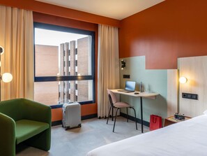 Hotellihuone, jossa on sänky, työpöytä kannettavalla tietokoneella, tuoli, matkalaukku ja punainen kassi.