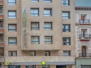Ein mehrstöckiges Hotel mit einheitlicher Fassade, zahlreichen Fenstern und einem grünen Schild, das auf ein Geschäft oder eine Dienstleistung hinweist.