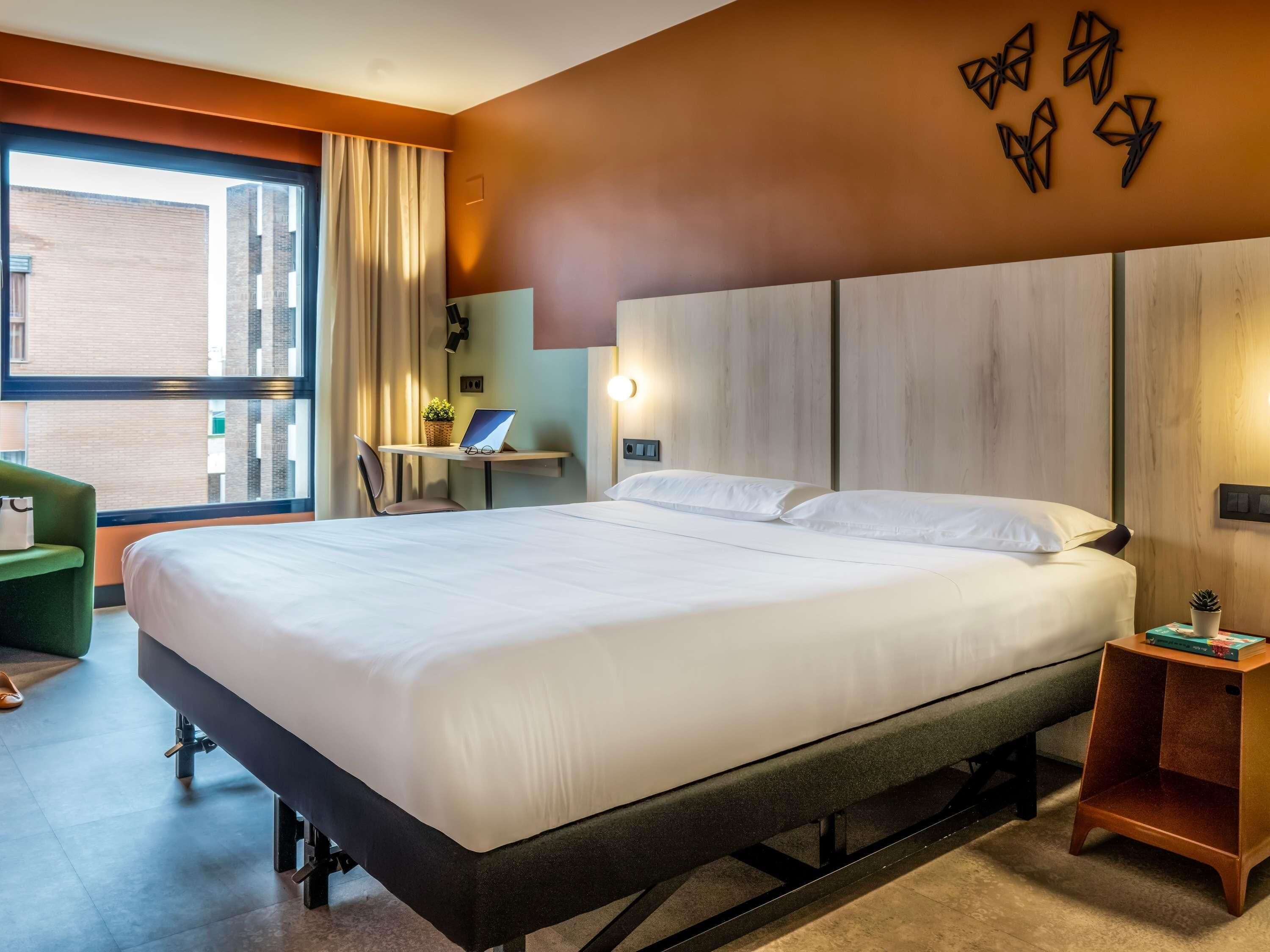 A hotel room with a bed, a desk with a laptop, a chair, a small table with books, and a window with curtains.