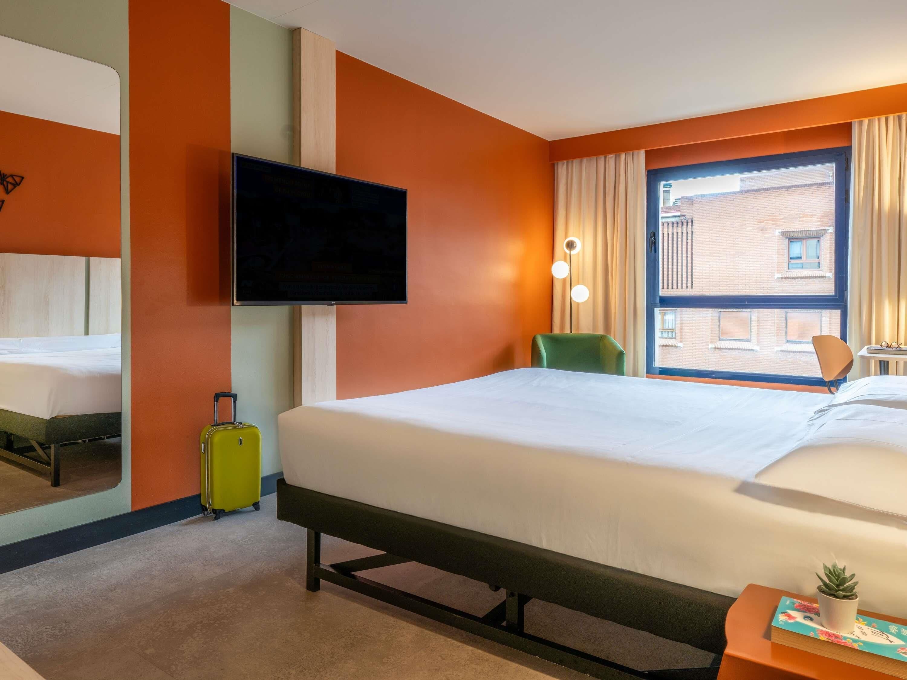 A hotel room with a bed, a television, a window with curtains, a chair, and a small table with a plant.