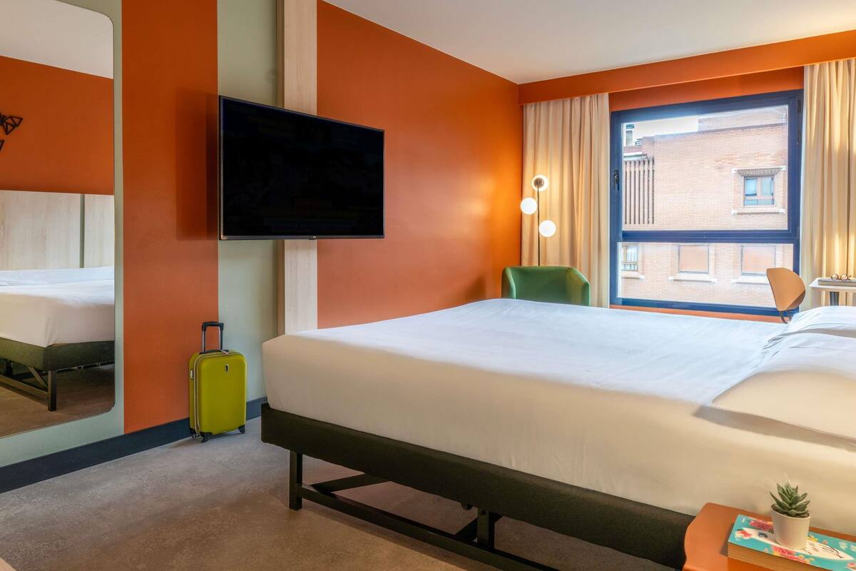 A hotel room with a bed, a television, a window with curtains, a chair, and a small table with a plant.