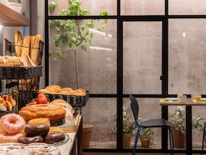 A bakery display with various baked goods, a window with a view of greenery, and a table with a chair. - ibis Styles Zaragoza Centro (Zaragoza)