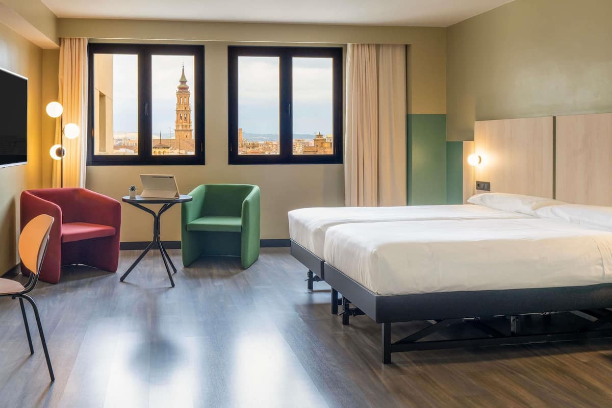 A hotel room with a bed, a desk with a laptop, a red chair, a green chair, and a view of a tower through large windows.