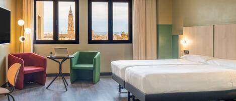 A hotel room with a bed, a desk with a laptop, a red chair, a green chair, and a view of a tower through large windows.