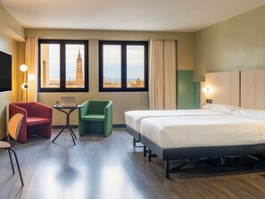 Hotellihuone, jossa on sänky, työpöytä kannettavalla tietokoneella, punainen tuoli, vihreä tuoli ja näkymä tornille suurista ikkunoista.