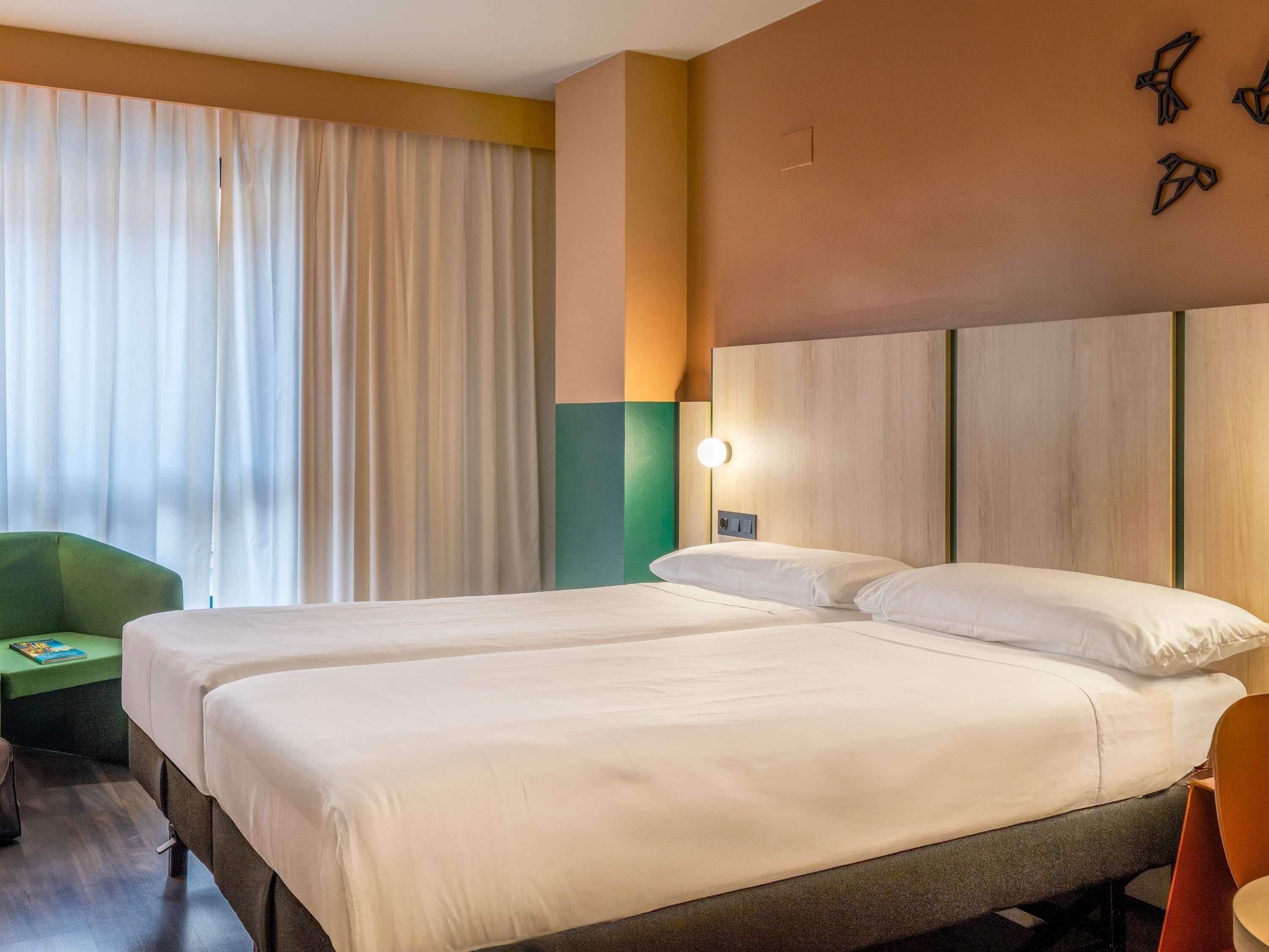 A hotel room with two beds, a green chair, and a wall-mounted light fixture.